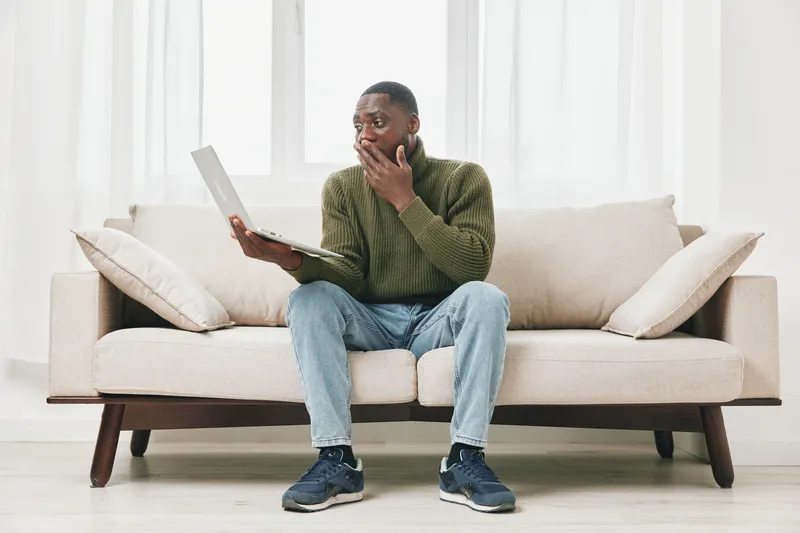 Concerned African man sitting on a sofa with a laptop, showing surprise and anxiety in a minimalist home setting with bright natural light