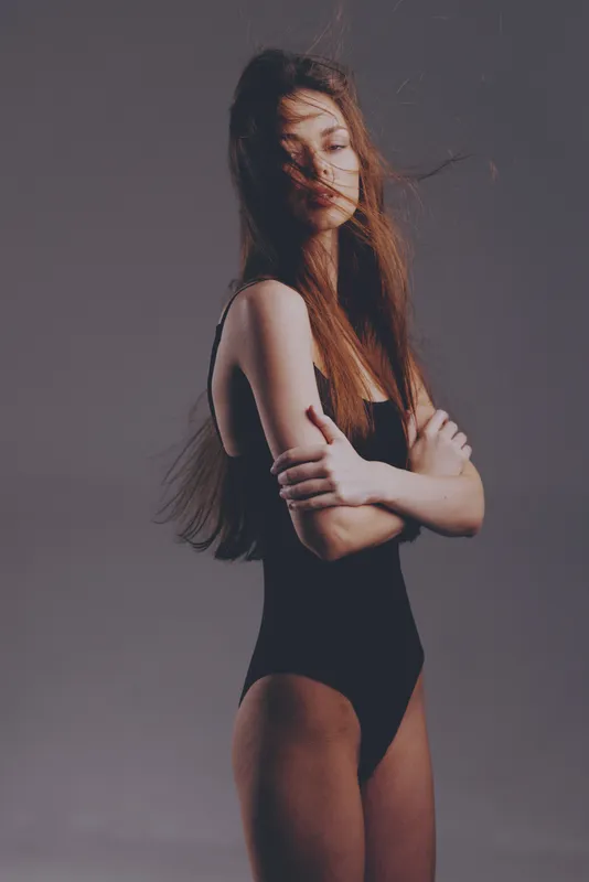 Young woman in black swimsuit posing with crossed arms and wind-blown hair on plain gray background.