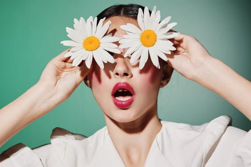 Creative beauty portrait of woman with glossy pink lips holding white daisies over eyes. Fashion concept with playful expression and pastel green background illuminating fresh unique style.