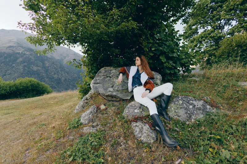 Woman lounging on rocks under a large tree in a mountain meadow, wearing stylish boots and a fashion jacket with fur cuffs, relaxed portrait capturing natural countryside serenity.