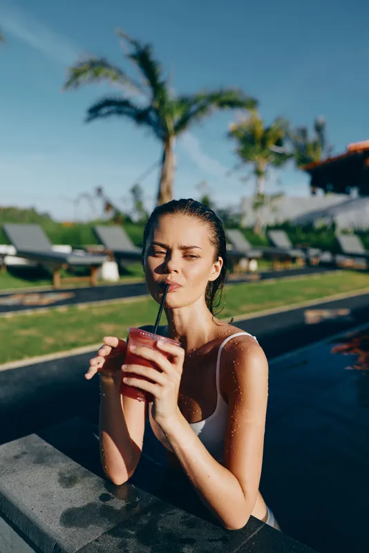 A woman in a light swimsuit relaxes in a pool, sipping a fruity drink. Bright outdoor scene with palm trees, blue sky, and sunlit water. Lifestyle moment capturing self care, leisure, and vacation