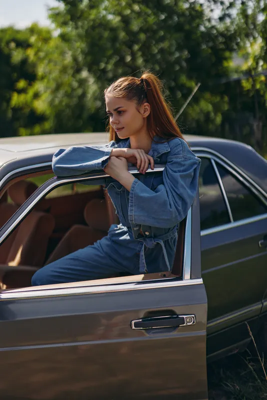 Young woman in denim attire poses stylishly in a vintage car outdoors, showcasing a casual and relaxed vibe with nature in the background.