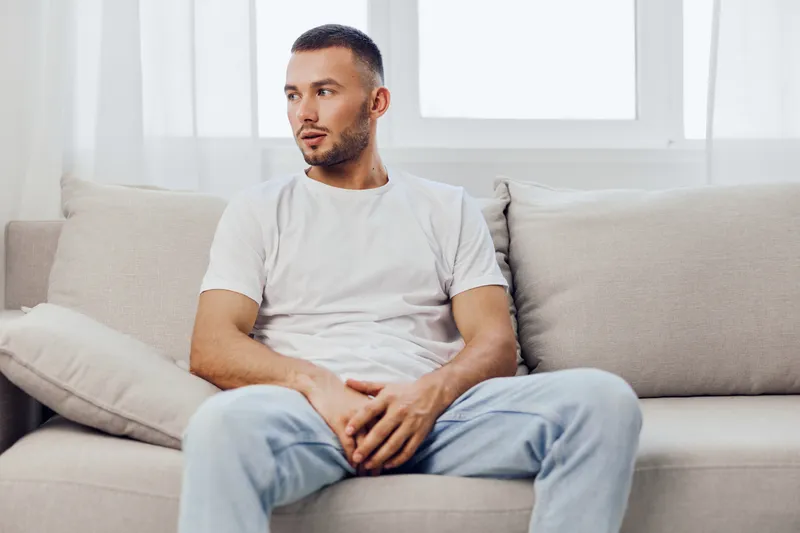 Thoughtful man sitting on a cozy couch, reflecting in a bright, modern living space. Natural light highlights his calm demeanor, evoking a sense of tranquility.
