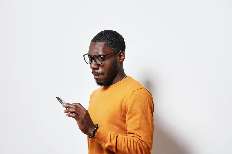 African American man with dark skin wearing glasses and orange sweater using smartphone for communication and social media on white background in casual style.