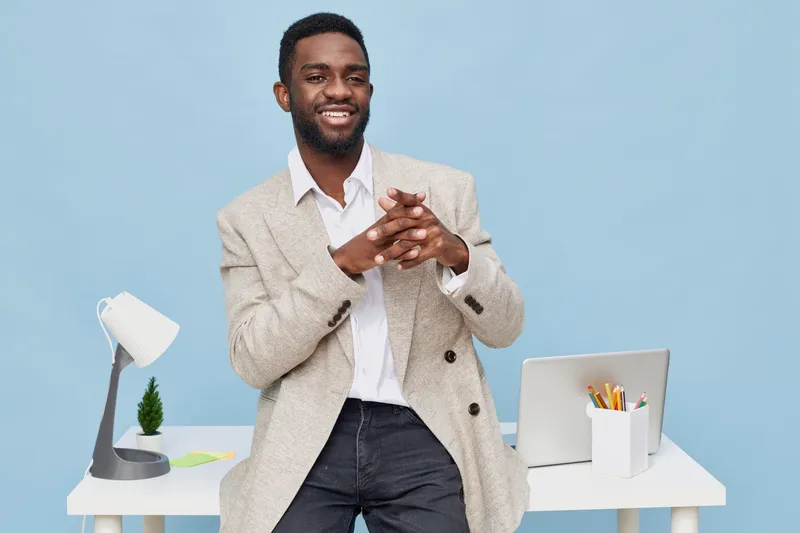 Smiling man dressed in a beige blazer and white shirt standing at a modern white desk, holding a pen and looking confident, with a laptop and office accessories in a bright, minimalistic workspace. Pe