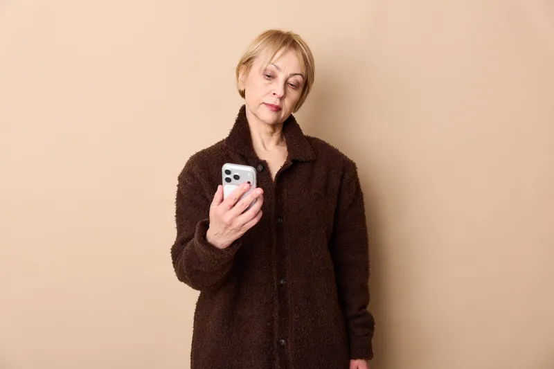 Senior woman holds a smartphone and phone, looking concerned and worried, with minimal communication and texting, concept for loneliness and stress indoors, beige background