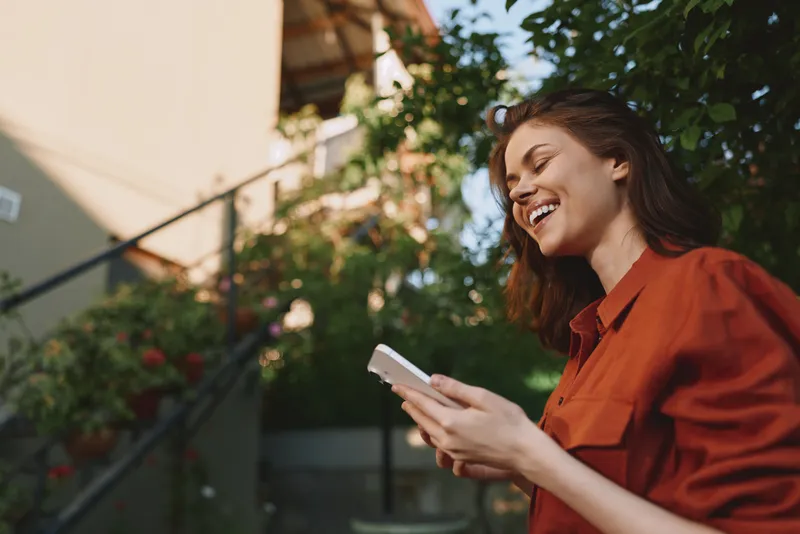 Cheerful woman outdoors, smiling while using a phone, sunlit garden setting, casual wardrobe, relaxed posture, natural colors, fresh air, vibrant greenery, candid lifestyle moment