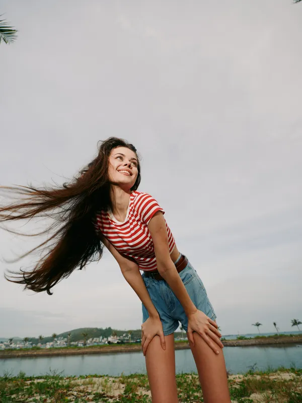 Joyful young woman bends slightly, wind tossing her hair, wearing a striped red and white shirt with denim shorts, a bright sky and water behind for a sunlit, carefree moment.