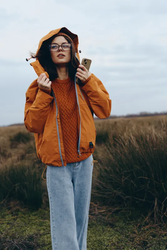 Woman lifestyle outdoors in a grassy field under wind, wearing an orange jacket and knitted scarf, round glasses, calm face, adjusting hood, natural daylight for travel and leisure