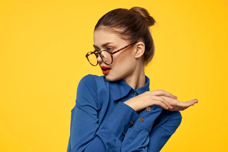 Женщина в синей рубашке и очках с ярким макияжем на лице жестикулирует руками на желтом фоне