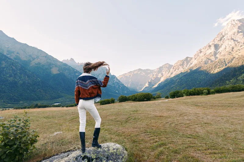 Woman standing in a mountains landscape meadow, traveler wearing a cozy sweater and boots on a rock, looking toward distant peaks and valley in serene outdoor scenery during golden hour.