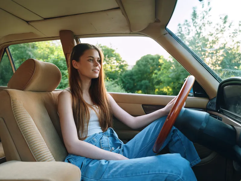 Young woman driving vintage car, wearing casual outfit with denim overalls, smiling and enjoying the ride in a lush green environment.