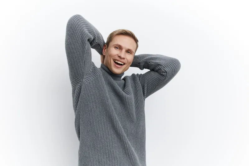 Happy smiling young man with blond hair wearing a cozy gray knitted sweater looking at camera exploring positive emotions against a plain white background studio portrait. People lifestyle concept