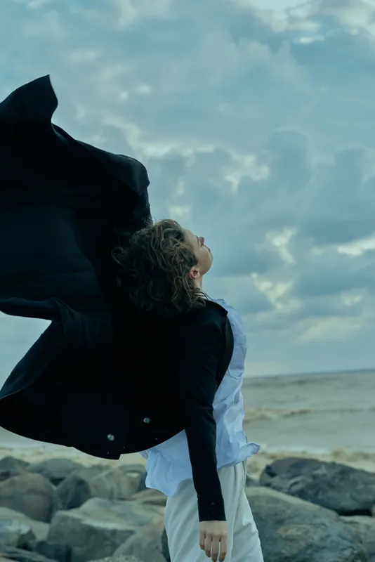 Wind sweeps a flowing cape as a figure stands on a windy beach, coat fluttering, showing movement and freedom against a moody sky and rocky shoreline