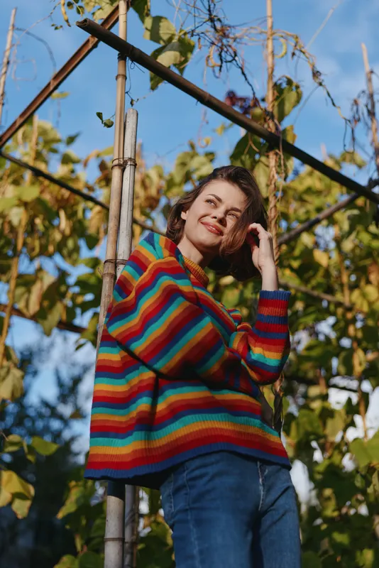 woman in rainbow sweater portrait in garden smiling and casual, leaning on trellis pole among vines, colorful autumn fashion, relaxed pose and natural light