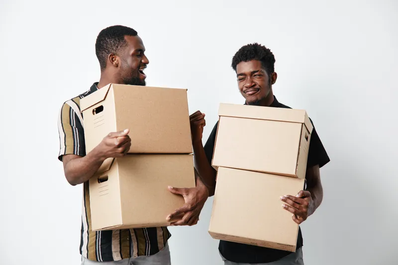 Two african american men with dark skin holding cardboard boxes for moving or delivery, smiling and standing on white background in casual clothes.