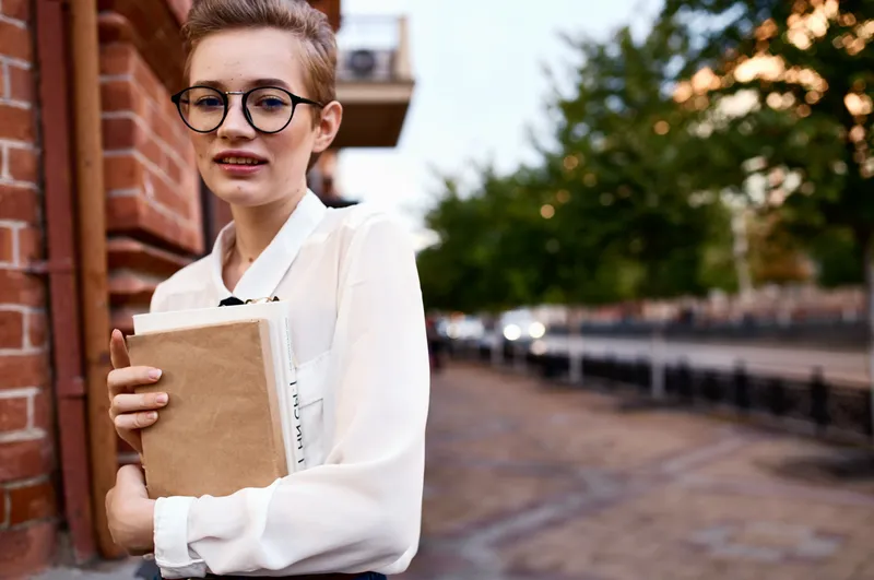 Студентка в очках с книгой стоит на улице возле института
