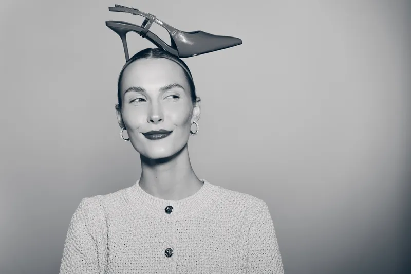 Young white woman with pale skin wearing a shoe on her head and a textured jacket looks sideways with a playful smile in a studio setting. Concept of fashion humor and creativity.