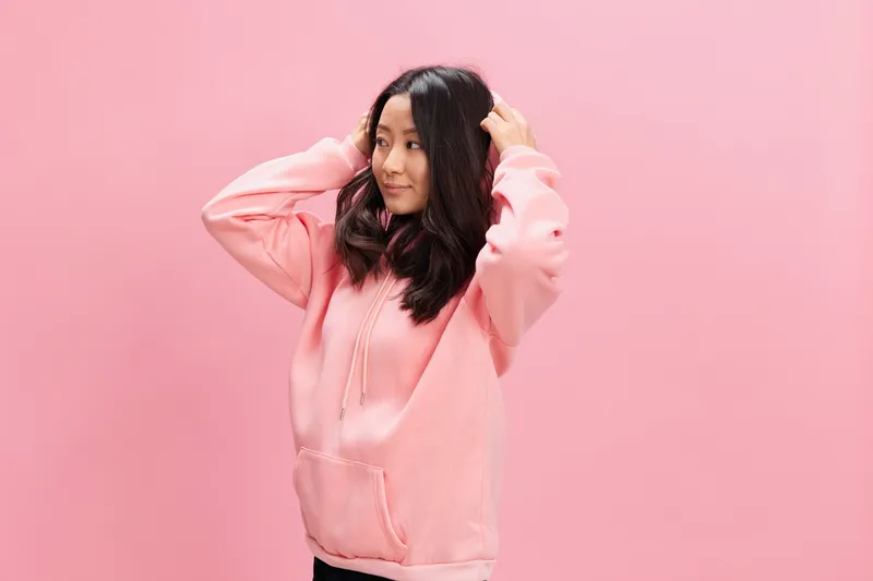 Вид сбоку: милая молодая азиатская студентка в розовой толстовке с капюшоном позирует изолированно на розовом студийном фоне