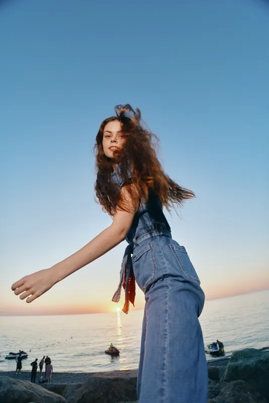 lifestyle woman in modern boho-western denim street style with loose hair at sunset by the sea, film color, casual and free-spirited atmosphere
