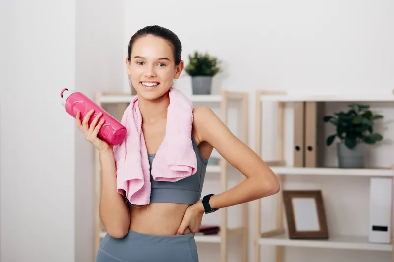Fitness woman in sportwear holds a pink water bottle with a towel around neck, standing in a clean home gym with shelves and plants, ready for workout.