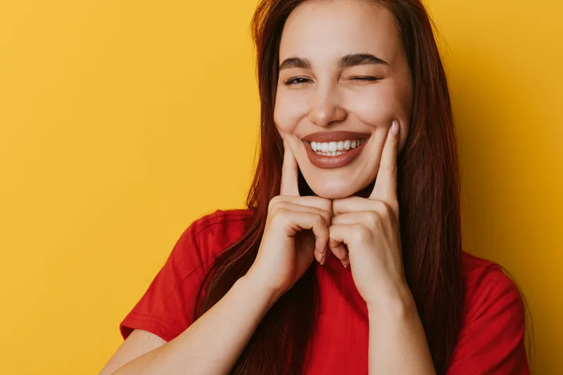 Woman with long hair wearing red shirt smiling and winking with fingers on cheeks against yellow background. Portrait of happy female showing positive expression and joy.