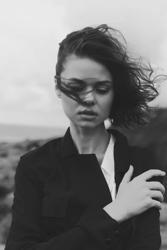 Outdoor shot of a woman in a windy setting, wearing a dark coat, moody expression, monochrome tones, raw natural textures