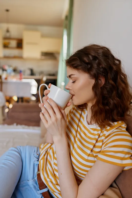 A relaxed woman sits at home, drinking from a mug, wearing a striped tee and jeans. Bright natural light spills over a cozy living space, creating a calm, intimate morning mood and everyday comfort.
