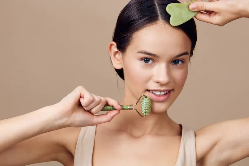 Young woman using a jade roller for skincare with a gua sha tool, showcasing glowing skin and a natural beauty routine in a minimalistic background