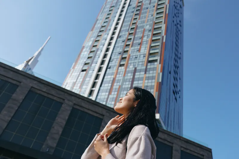 Woman looking up at a towering skyscraper in a sunny urban setting, highlighting ambition, architecture, and city life beneath a clear blue sky with glass facades and modern streets in stock