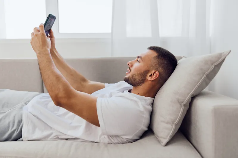 Relaxed man lying on the couch, enjoying the latest app on his smartphone while surrounded by a minimalist and bright living space.