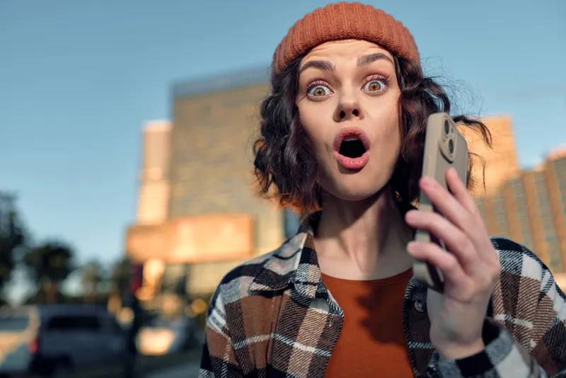 Woman with phone shows surprised expression in urban portrait during golden hour, candid lifestyle moment with authenticity, emotional storytelling, mindful living and warm glow.