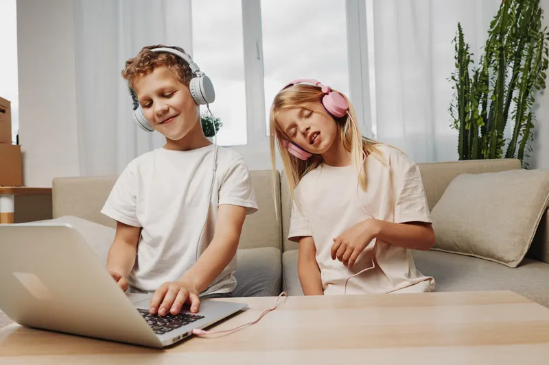 Happy boy and girl wearing headphones enjoy music together at home, sitting on couch with laptop on table. Concept of childhood fun, technology, and shared entertainment indoors.