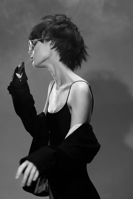Black and white portrait of a stylish woman with short hair wearing sunglasses and a loose jacket in a moody atmosphere.
