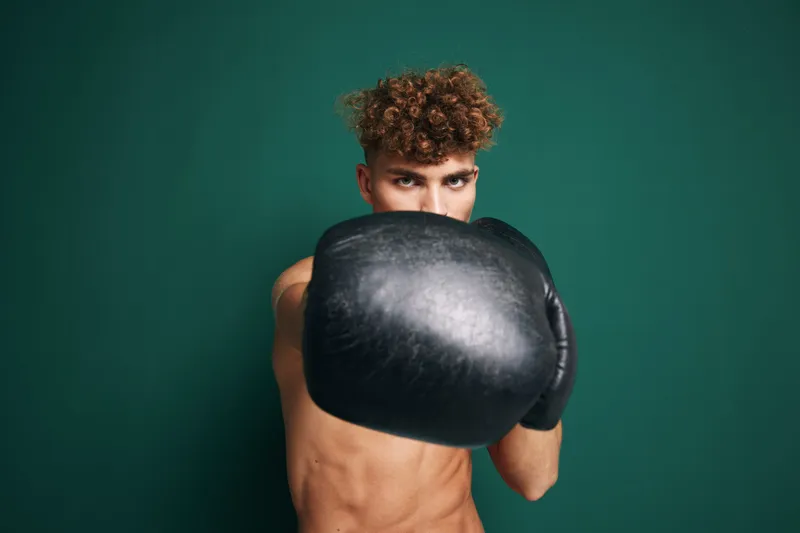 Young man with curly hair wearing black boxing gloves in fighting stance against green background. Athlete training, fitness and combat sport concept with focus on strength and determination.