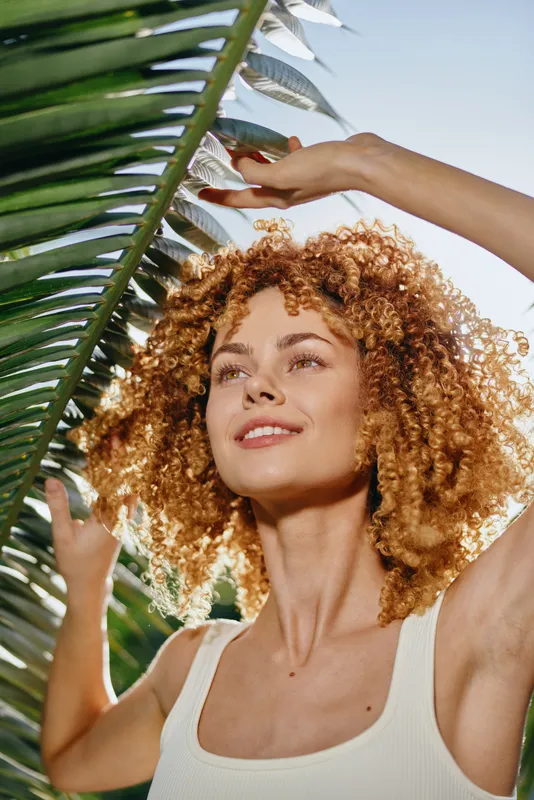smiling woman with curly hair enjoying sunlight outdoors near tropical palm leaves, wearing white tank top under clear blue sky during summer day