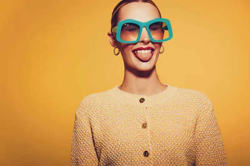 Young woman in large blue sunglasses and beige sweater sticks out tongue against yellow background. Fun, playful mood with bright colors and casual fashion style.
