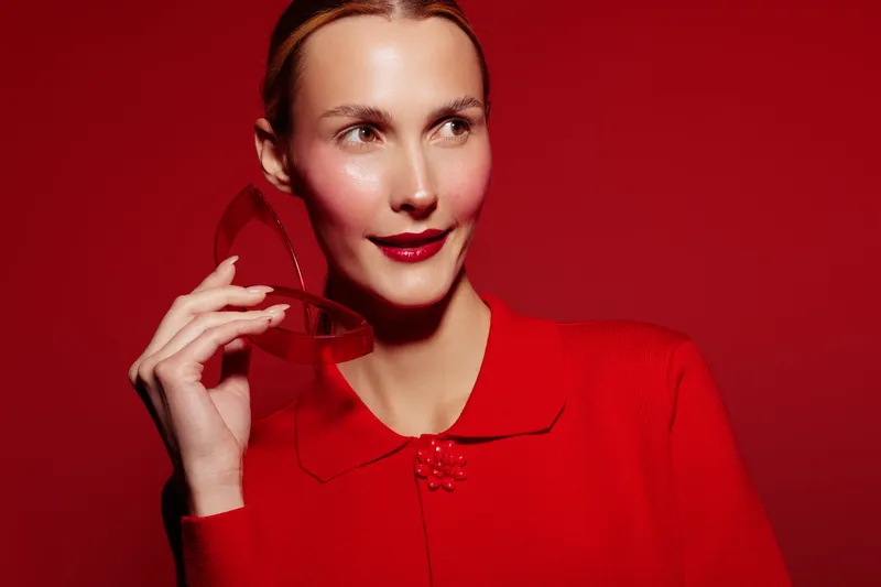 Young woman in red sweater holds red sunglasses near face, looking aside with slight smile. Bright red background creates bold and stylish fashion portrait concept.
