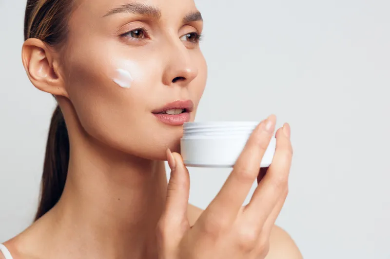 Young woman applies moisturizing cream on face holds white jar skincare beauty routine natural fresh skin care isolated on light background closeup portrait
