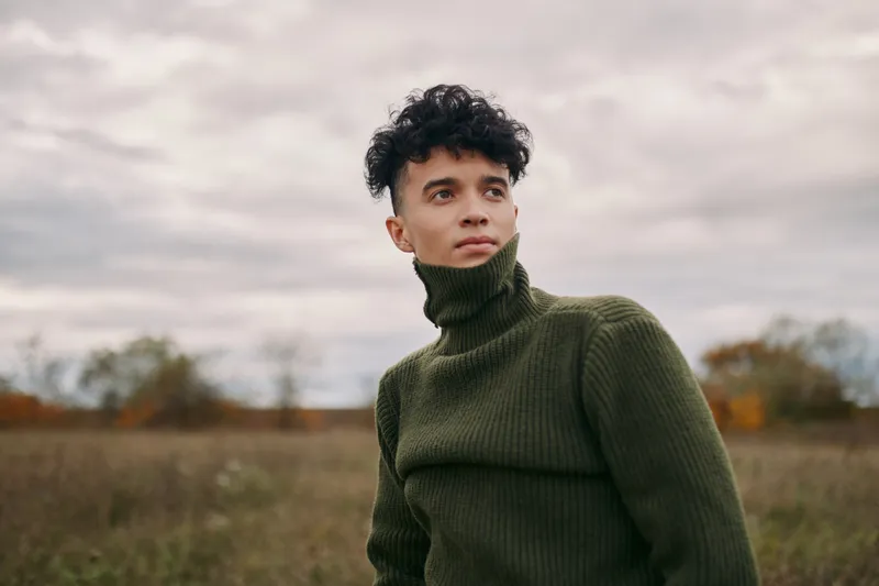 Young man in an olive turtleneck sweater gazes across a quiet field, a genuine unposed portrait that highlights realism, natural light and subtle personal emotion.
