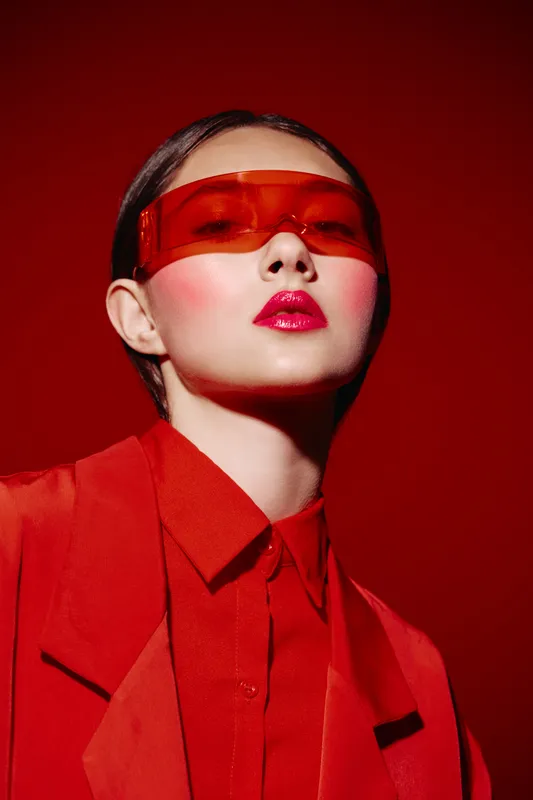 Young woman in bold red blazer and shirt poses confidently with oversized red tinted sunglasses on monochrome red background. Concept of beauty fashion and creative style.