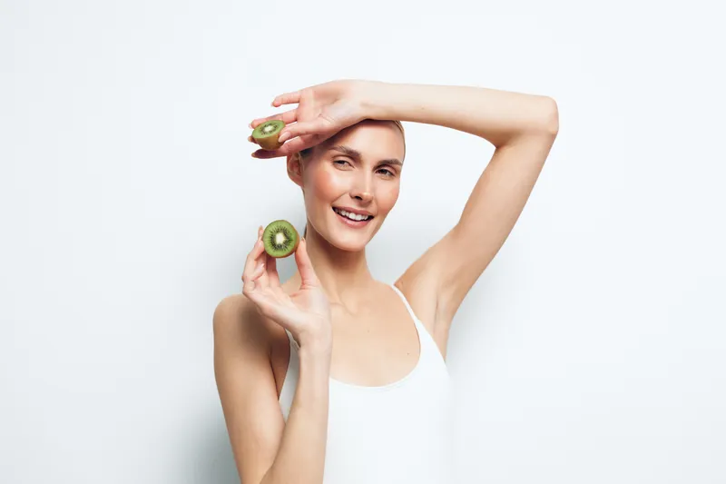 Smiling woman holding two kiwi halves near face on white background. Concept of healthy lifestyle, natural food, wellness, and fresh fruit. Soft lighting creates bright and clean mood.
