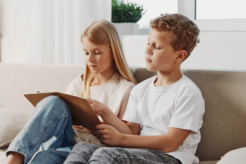 Two focused children sitting on sofa reading a book together in bright living room. Concept for learning, childhood, education, and family bonding.