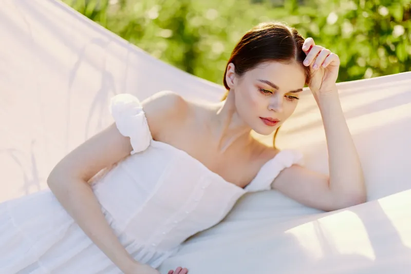 A serene young woman in a light summer dress, resting in a hammock amidst lush greenery, conveying tranquility and warmth during a sunny day