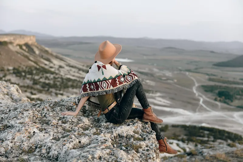 Женщина отдыхает на камне, на ней шляпа, а вокруг неё обернуто одеяло