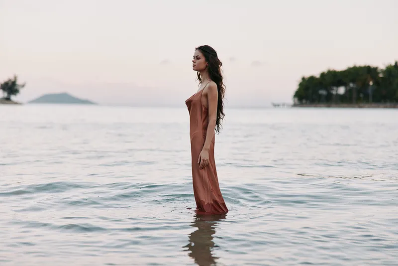A solitary woman stands waist-deep in calm ocean water, wearing a flowing terracotta dress as soft light brushes the horizon and gentle waves frame a serene moment.