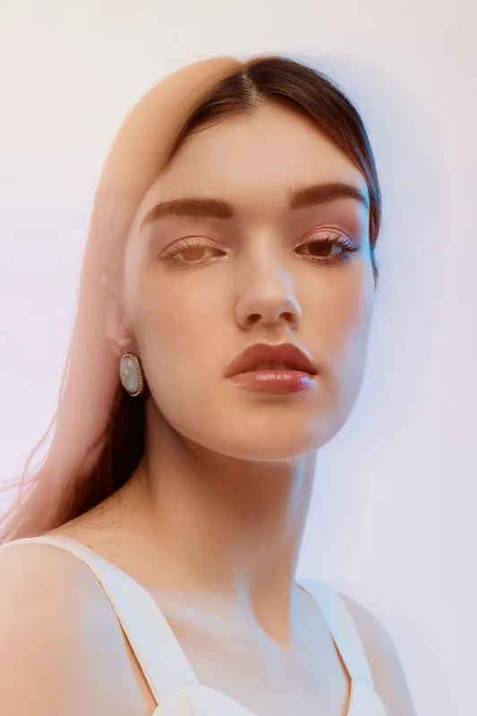 Close-up portrait of a young woman with natural makeup and glossy lips wearing white earrings and a white top. Soft lighting highlights her beauty and fashion style.