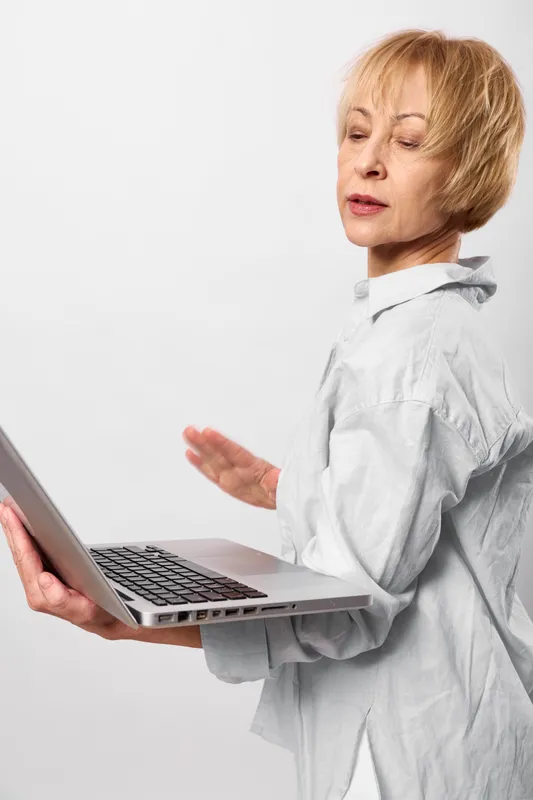 Elderly woman with laptop shows stress and concern, one hand gesturing while looking away, remote communication concept on plain white background, office technology support, today, care
