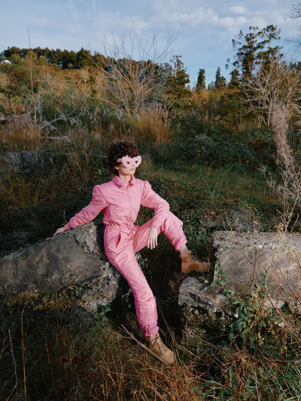 Pink jumpsuit wearing person sits on rocky terrain in an outdoor scene, surrounded by autumnal trees and shrubbery, posing confidently amid rugged natural landscape.
