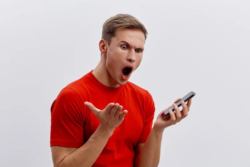 Excited young man in bright red t-shirt reacting with surprise or shock while looking at smartphone on plain gray background studio portrait. People emotion concept, surprised, communication, technolo